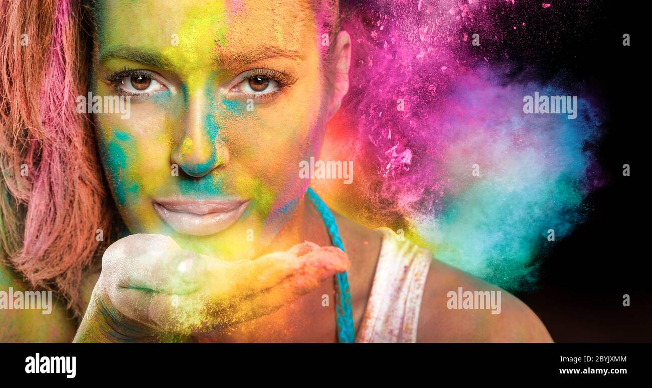 Beautiful young woman celebrating Holi fest with colorful powder covering her face and blowing a cloud of dust off the palm of her hand at the camera. Stock Photo