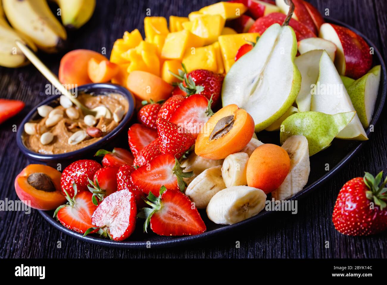 Rainbow fruit platter of fresh fruits and berries with peanut dip: strawberries, tropical mango, banana, apples, pears, apricots and peanuts on a blac Stock Photo