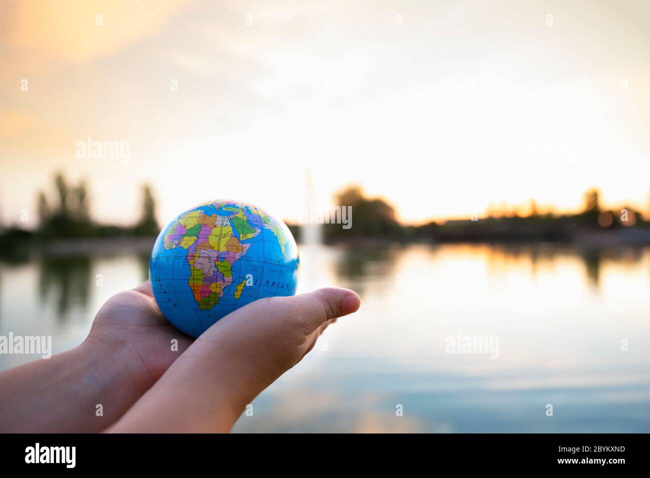 Detail of person's hands holding small ball of the world in front of a lake at sunset. Selective focus. Concept of travel and care of the planet. Stock Photo