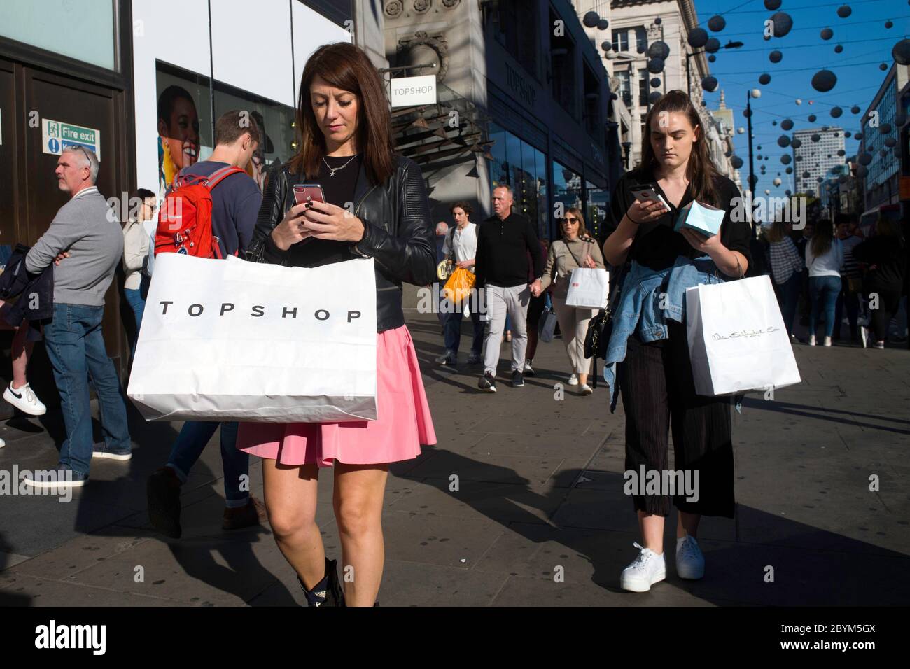 Two young women on their mobile phones walking down Oxford Street London carrying Top Shop shopping bags. Stock Photo