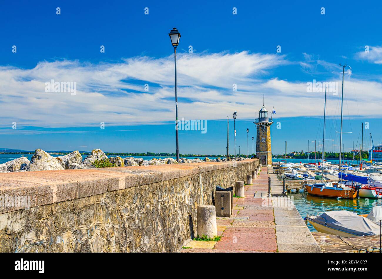 Stone pier mole with lighthouse, street lights and yachts on boat parking port marina in Desenzano del Garda town, blue sky white clouds background, Lombardy, Northern Italy Stock Photo