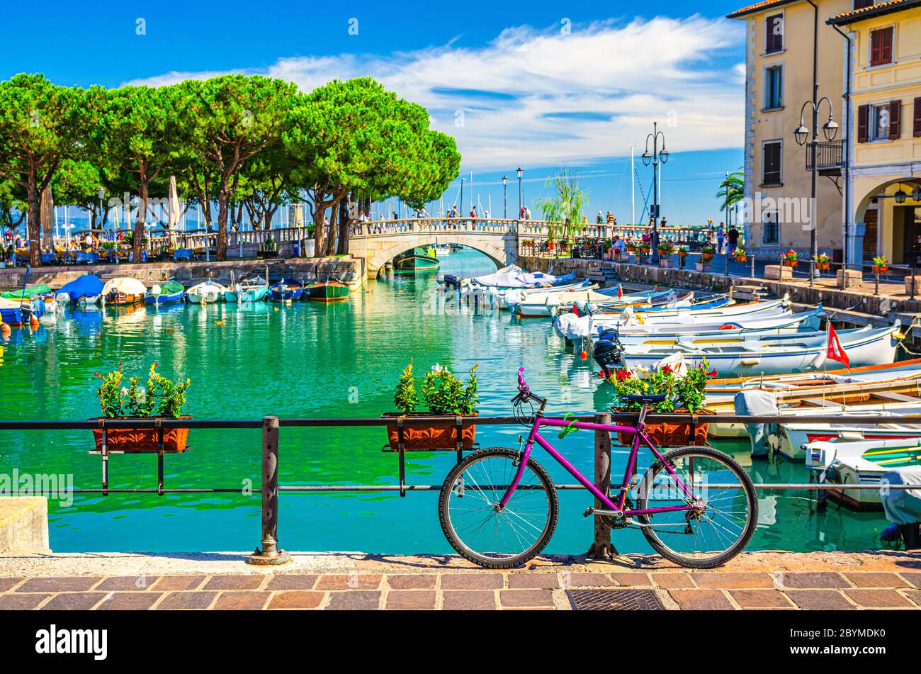 Bicycle bike near fence of old harbour Porto Vecchio with motor boats on turquoise water and Venetian bridge in historical centre of Desenzano del Garda town, blue sky, Lombardy, Northern Italy Stock Photo