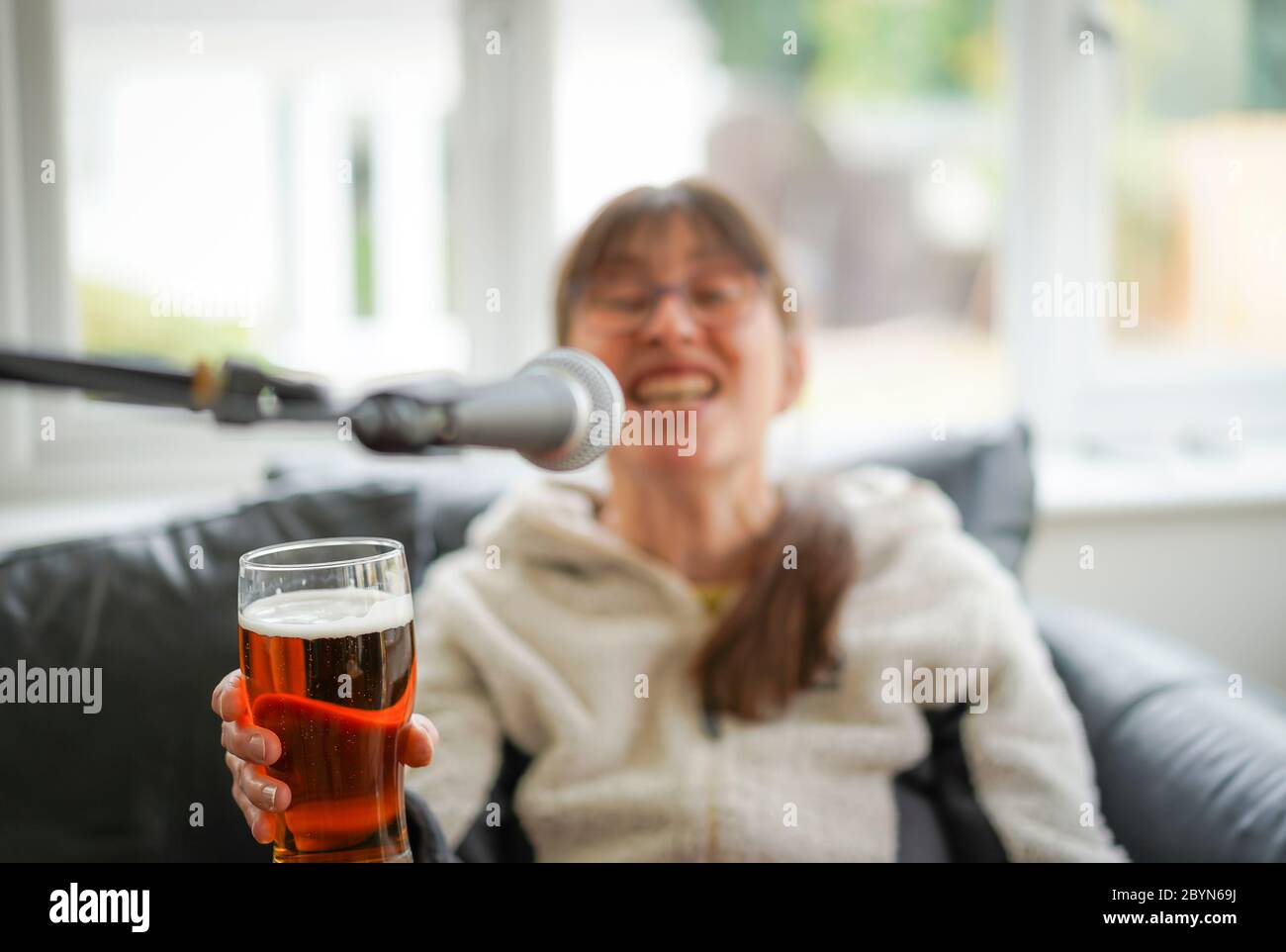 Isolated young woman drinking beer at home and singing into microphone. Drunk woman holding a pint of beer & enjoying herself! Drinking excess alcohol. Stock Photo