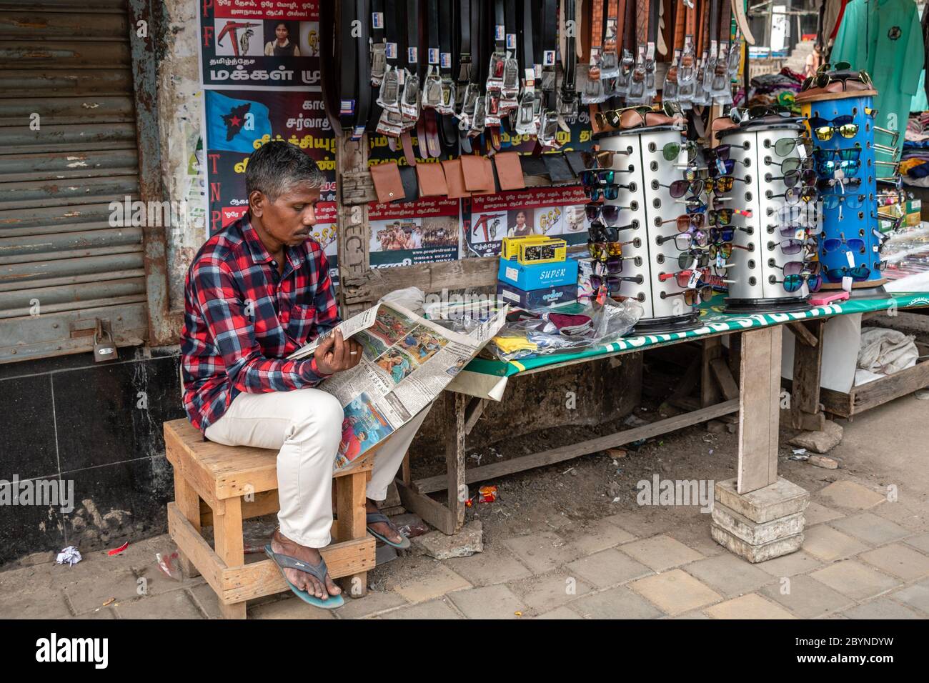 Chennai, Tamil Nadu, India - August 2018: A street vendor reading a newspaper outside his pavement shop that sells leather goods and sunglasses. Stock Photo
