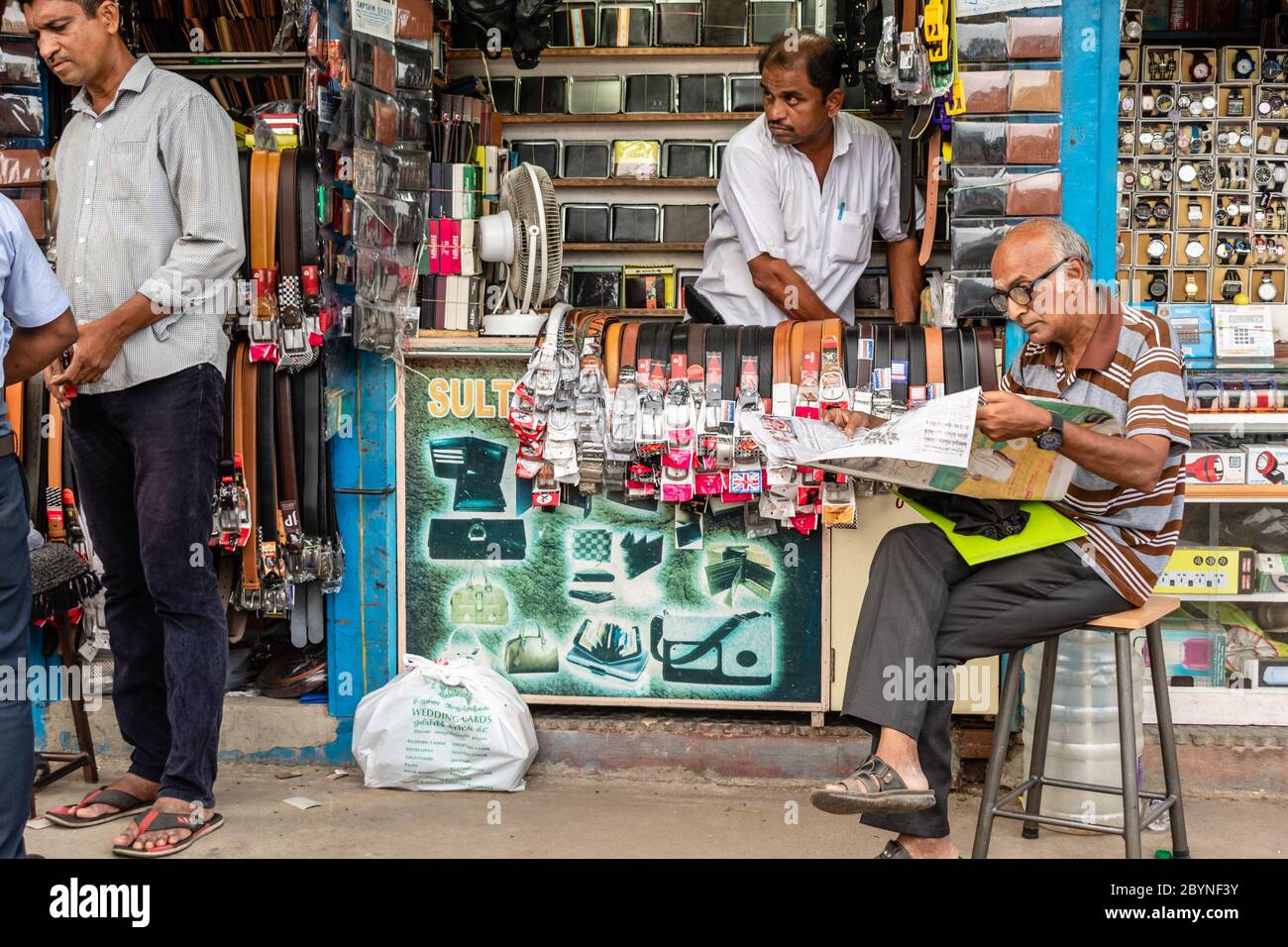 Chennai, Tamil Nadu, India - August 2018: An elderly Indian man reads a newspaper sitting outside a shop that sells belts and leather goods. Stock Photo