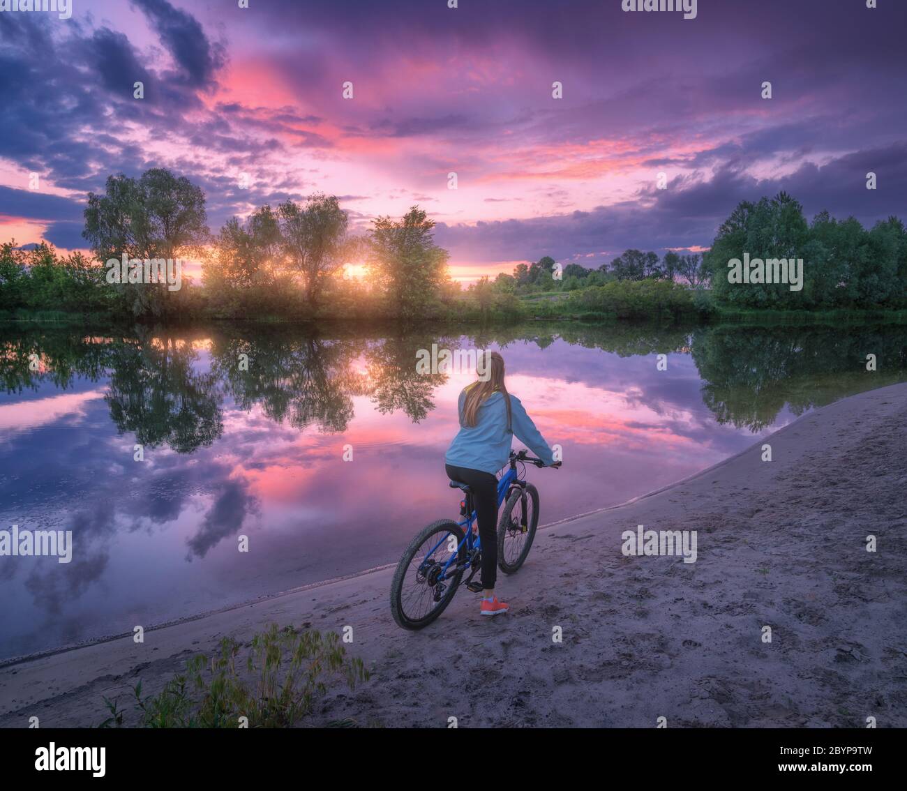 Woman with bicycle on the coast of the river at sunset in summer Stock Photo
