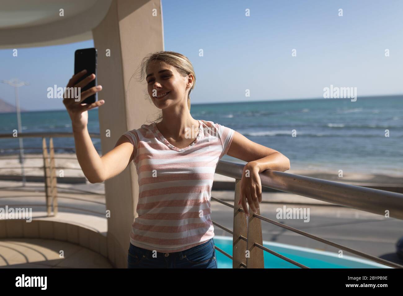 Caucasian woman standing on a balcony holding a smartphone and taking a selfie Stock Photo