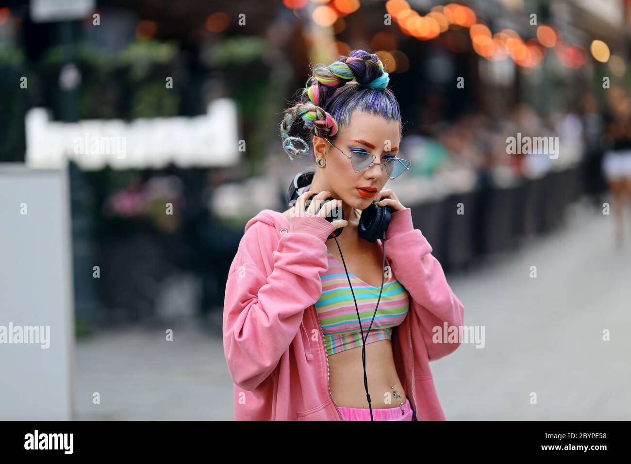 Fashion cool funky girl in headphones listening to music wearing colorful pink sweater and fancy sunglasses on the streets with pubs and bars in city Stock Photo