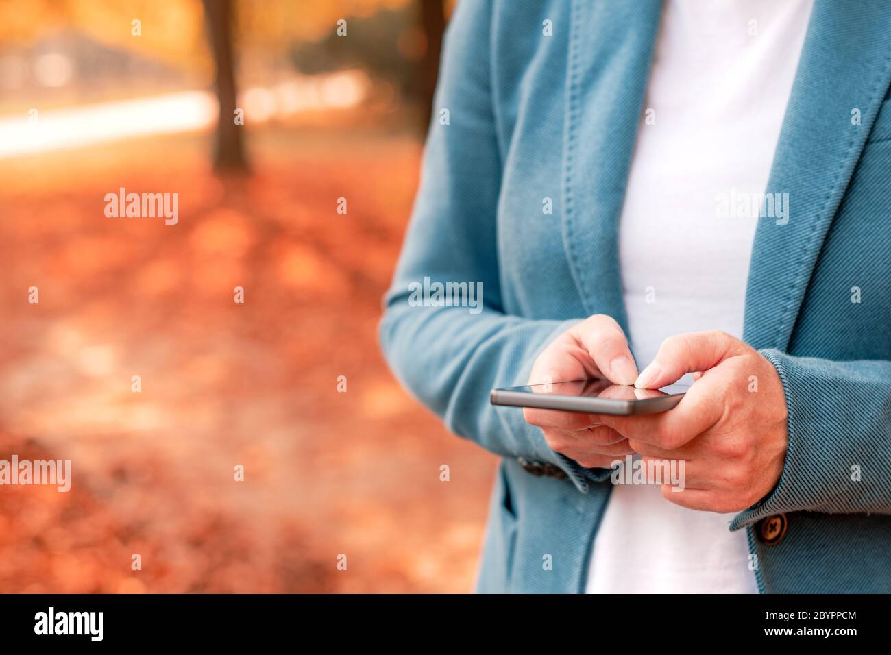 Businesswoman typing text message on smartphone in autumn park, selective focus on fingers Stock Photo