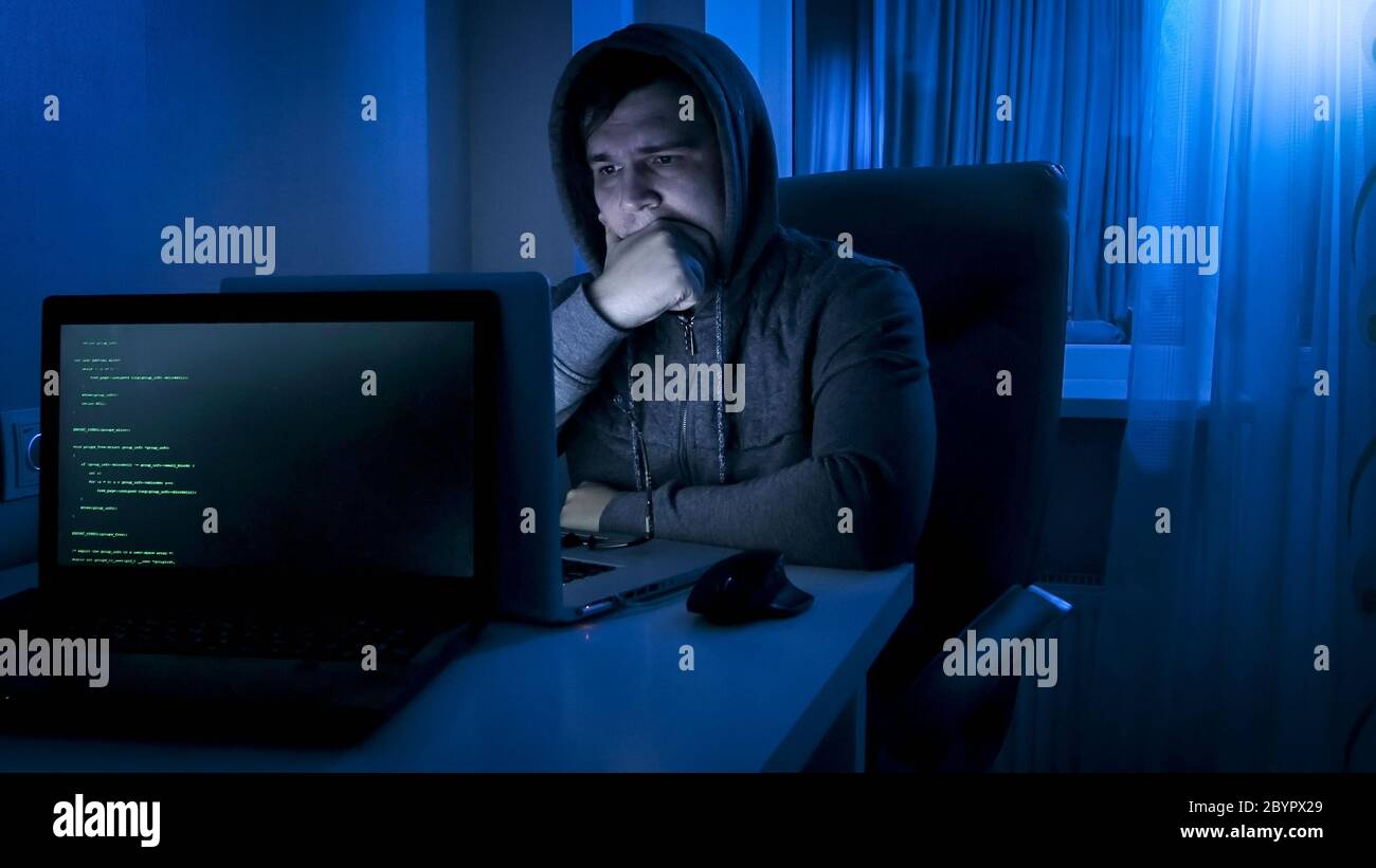 Thoughtful programmer in hood looking on laptop screen while working at night Stock Photo