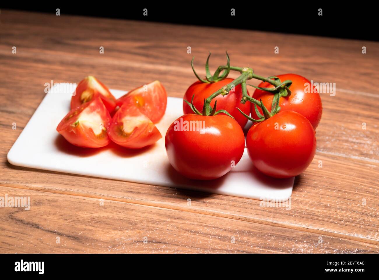 Ripe tomato chopped into four pieces with whole tomatoes on the right side on white chopping board wood background Stock Photo
