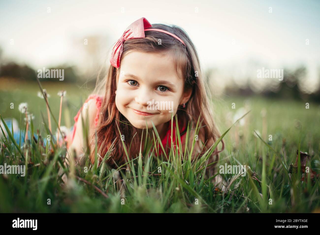 Cute adorable Caucasian girl among dandelions flowers. Child lying in grass on meadow. Outdoor fun summer seasonal children activity. Kid having fun o Stock Photo