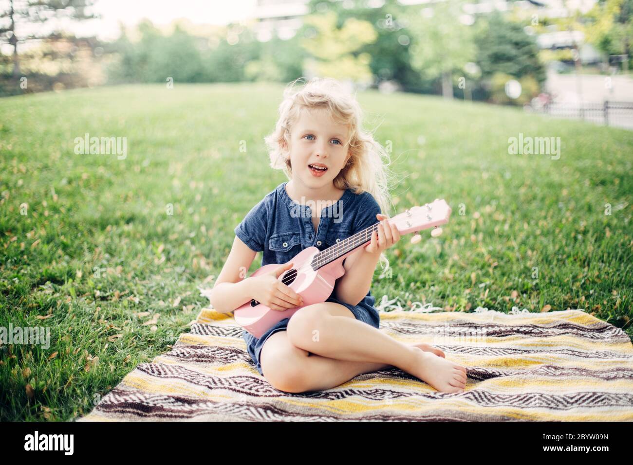 Cute adorable blonde girl playing pink guitar toy outdoor. Child playing music and singing song in park. Hobby activity for children kids. Tender memo Stock Photo