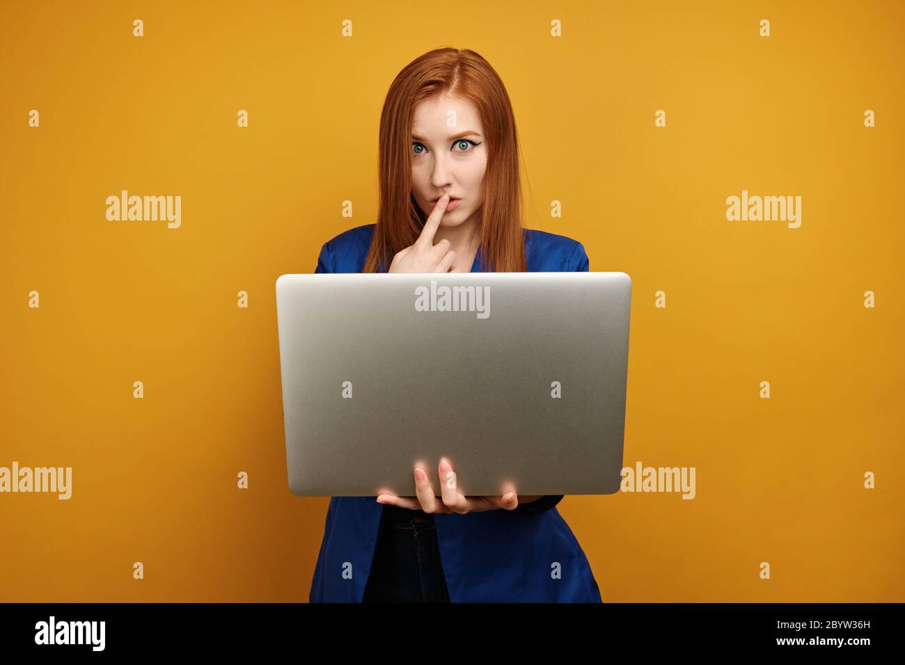 A red-haired girl in a blue jacket stands on a yellow background with a laptop in hand and putting finger to lips Stock Photo