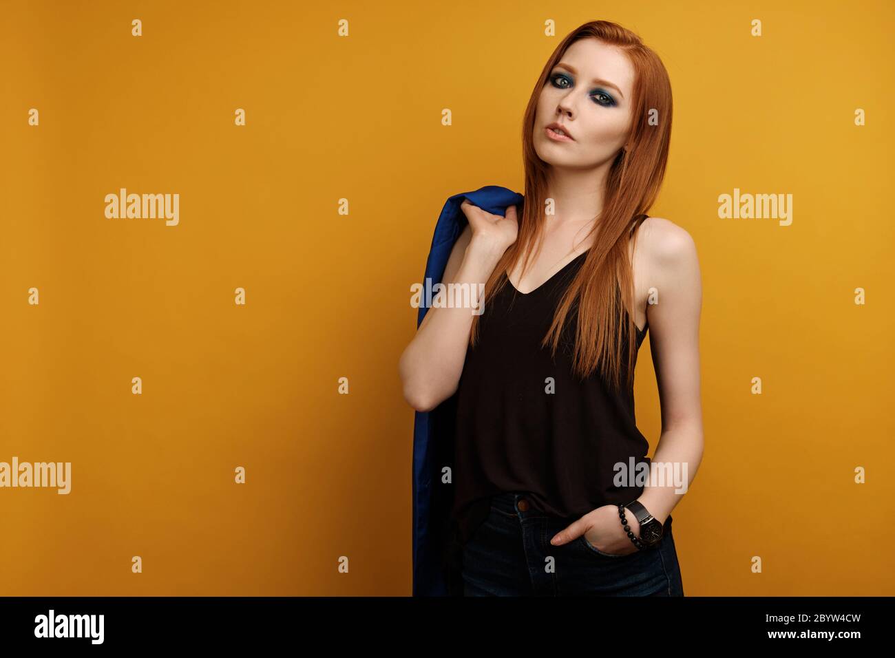 girl in a black top and blue smokey stands on a yellow background, throwing a jacket over shoulder and putting hand in pocket Stock Photo