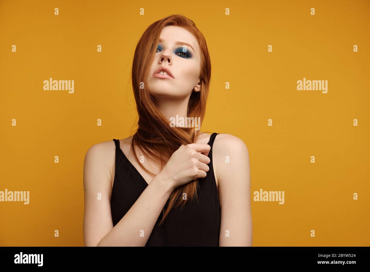 Redhead girl in a black top and with blue eye makeup stands on a yellow background with head thrown back and covered with hairs Stock Photo