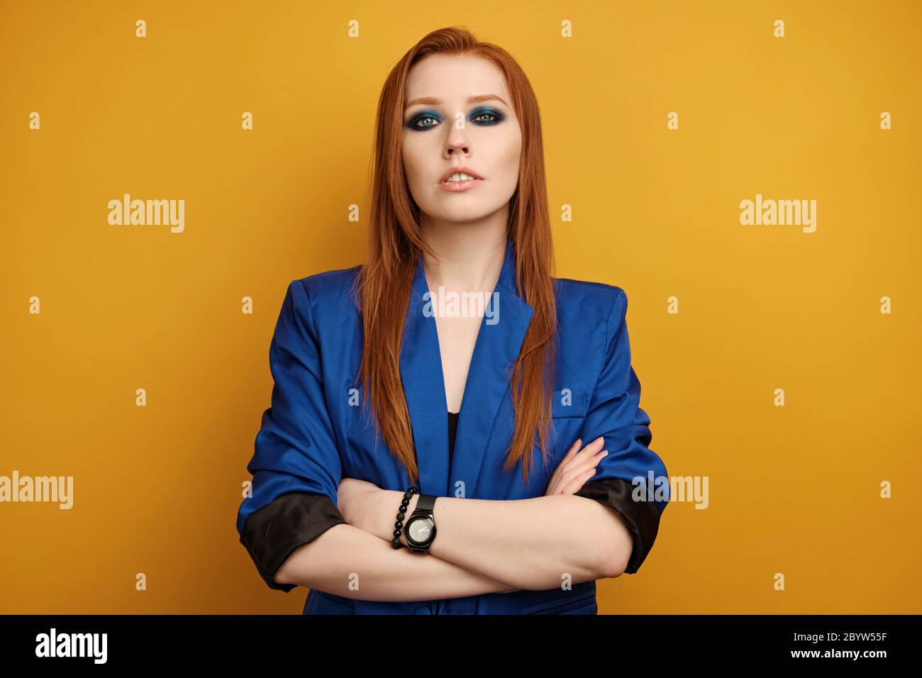 Red-haired girl in a blue jacket and with blue smokey stands on a yellow background with her arms crossed Stock Photo