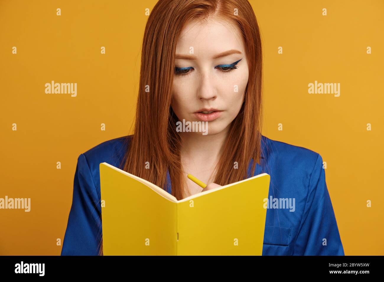 Red-haired girl in a blue jacket and with bright arrows stands on a yellow background and writes in a yellow notebook Stock Photo
