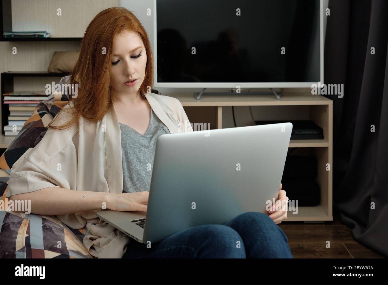 Red-haired girl sitting in a chair at home looking at a laptop on her lap Stock Photo