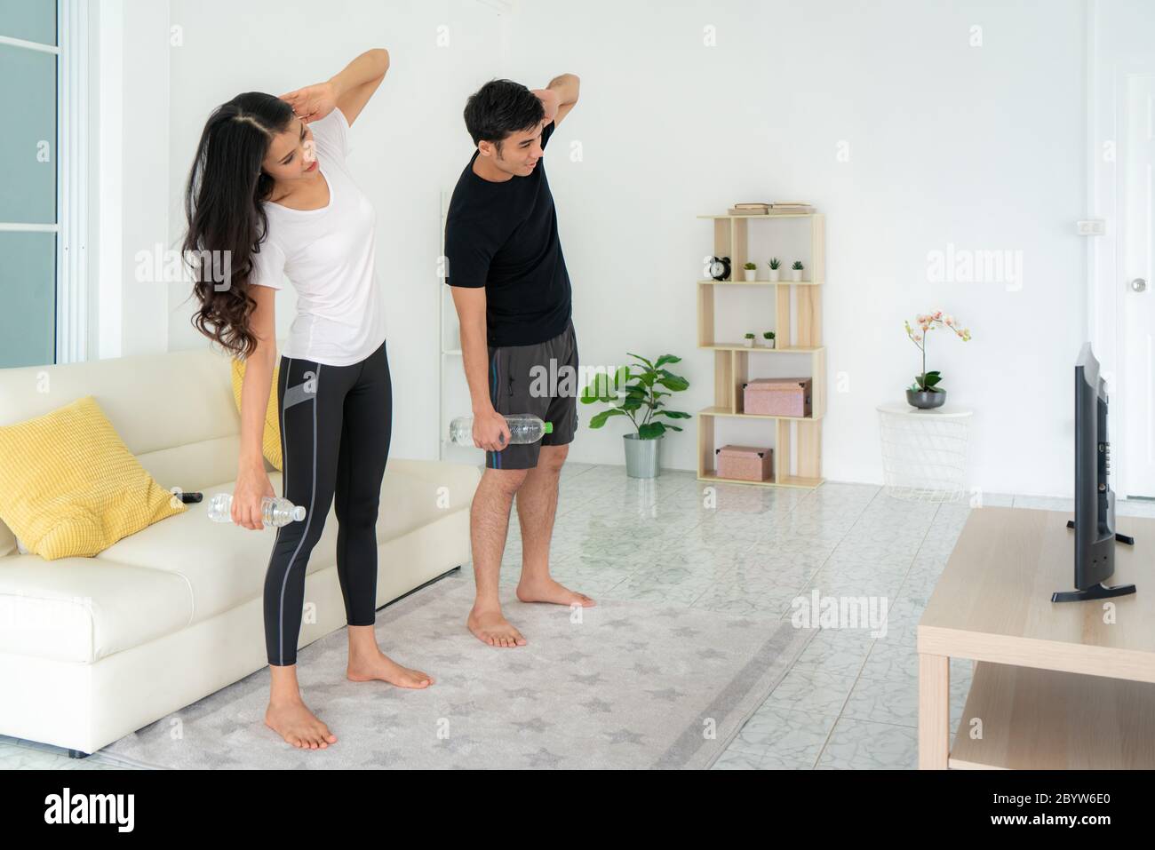 Young Asian couple doing High-intensity interval training together and looking TV at home, man and woman working out together standing in living room, Stock Photo