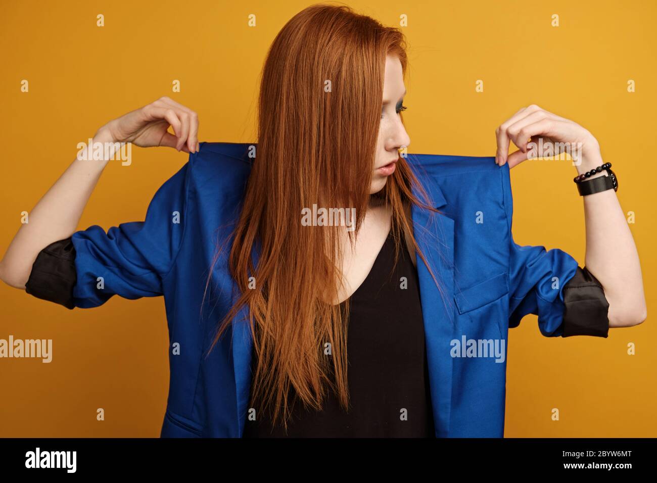 Redhead girl in a black top and a blue jacket stands on a yellow background holds the shoulders of the jacket with his fingers Stock Photo