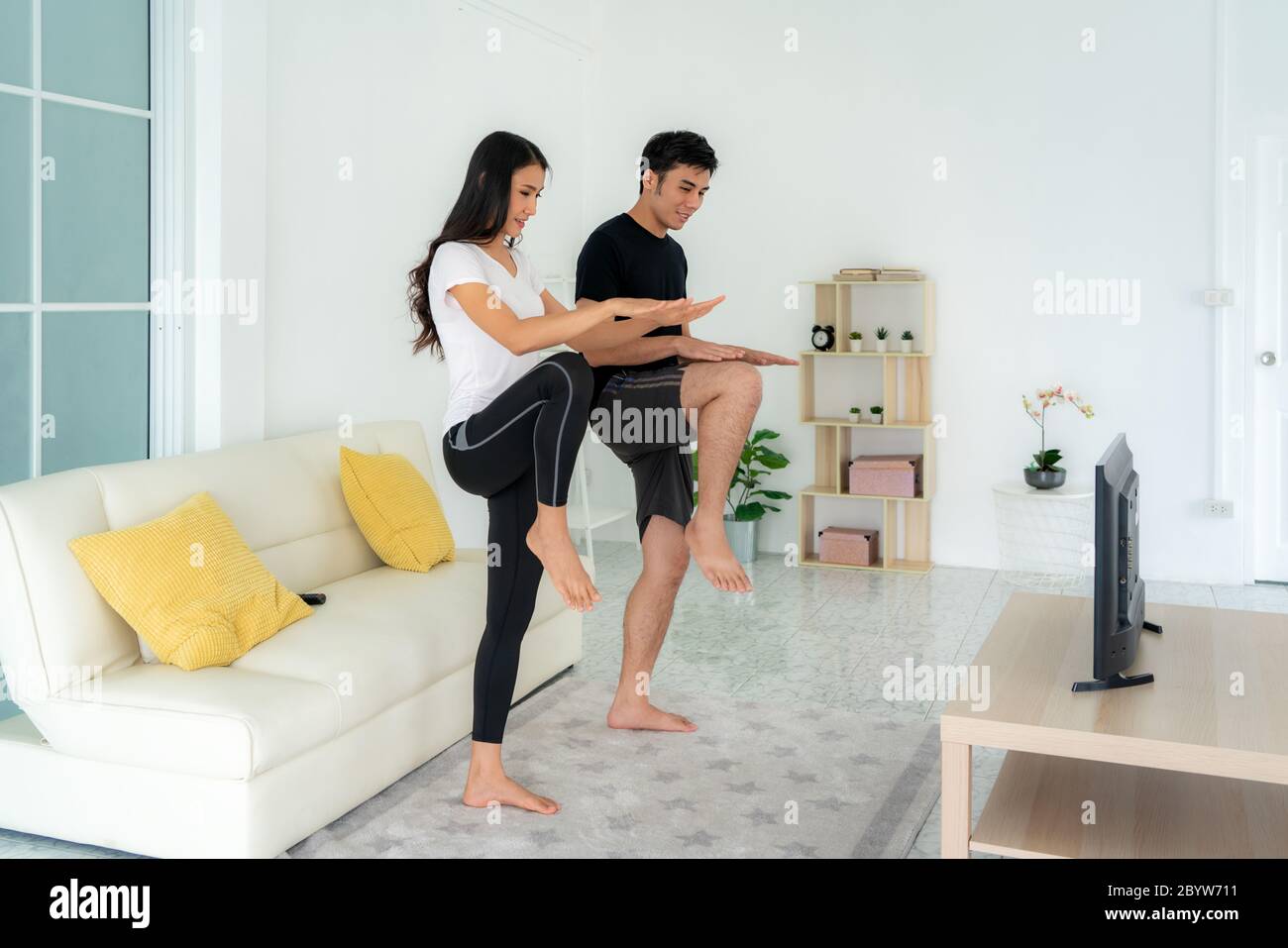 Young Asian couple doing High-intensity interval training together and looking TV at home, man and woman working out together standing in living room, Stock Photo
