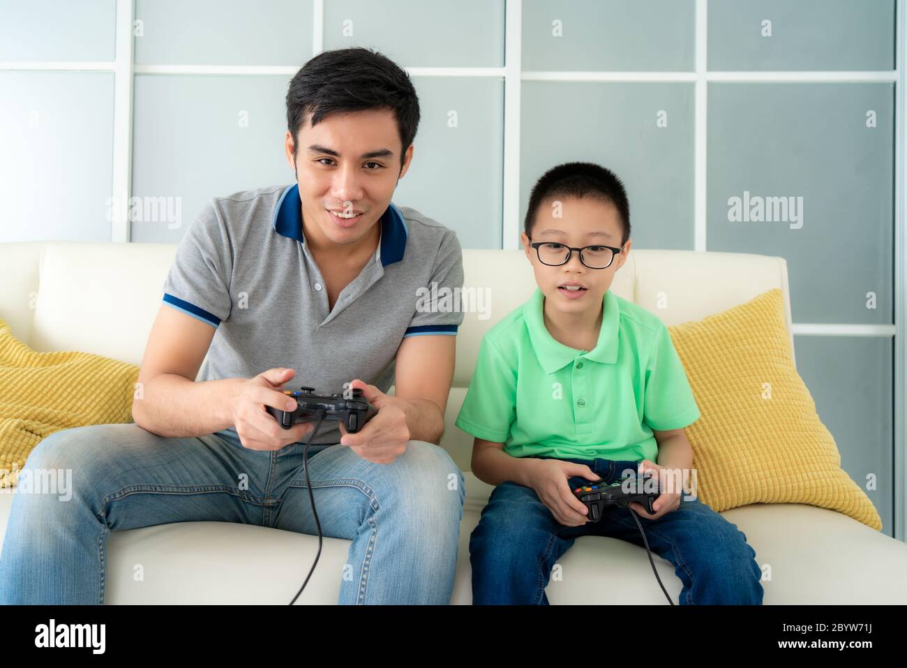 Asian family father and his son to playing video games with joysticks while sitting in sofa in living room at home, concept of family values and the m Stock Photo