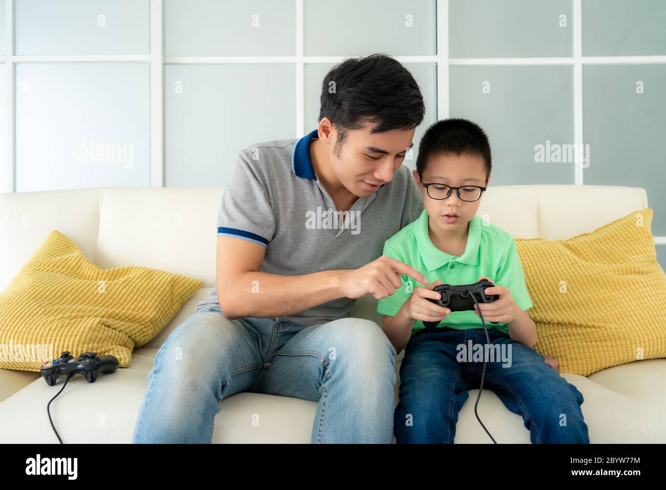 Asian family father teaching his son to playing video games with joysticks while sitting in sofa in living room at home, concept of family values and Stock Photo