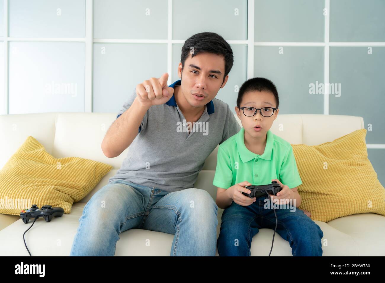 Asian family father teaching his son to playing video games with joysticks while sitting in sofa in living room at home, concept of family values and Stock Photo
