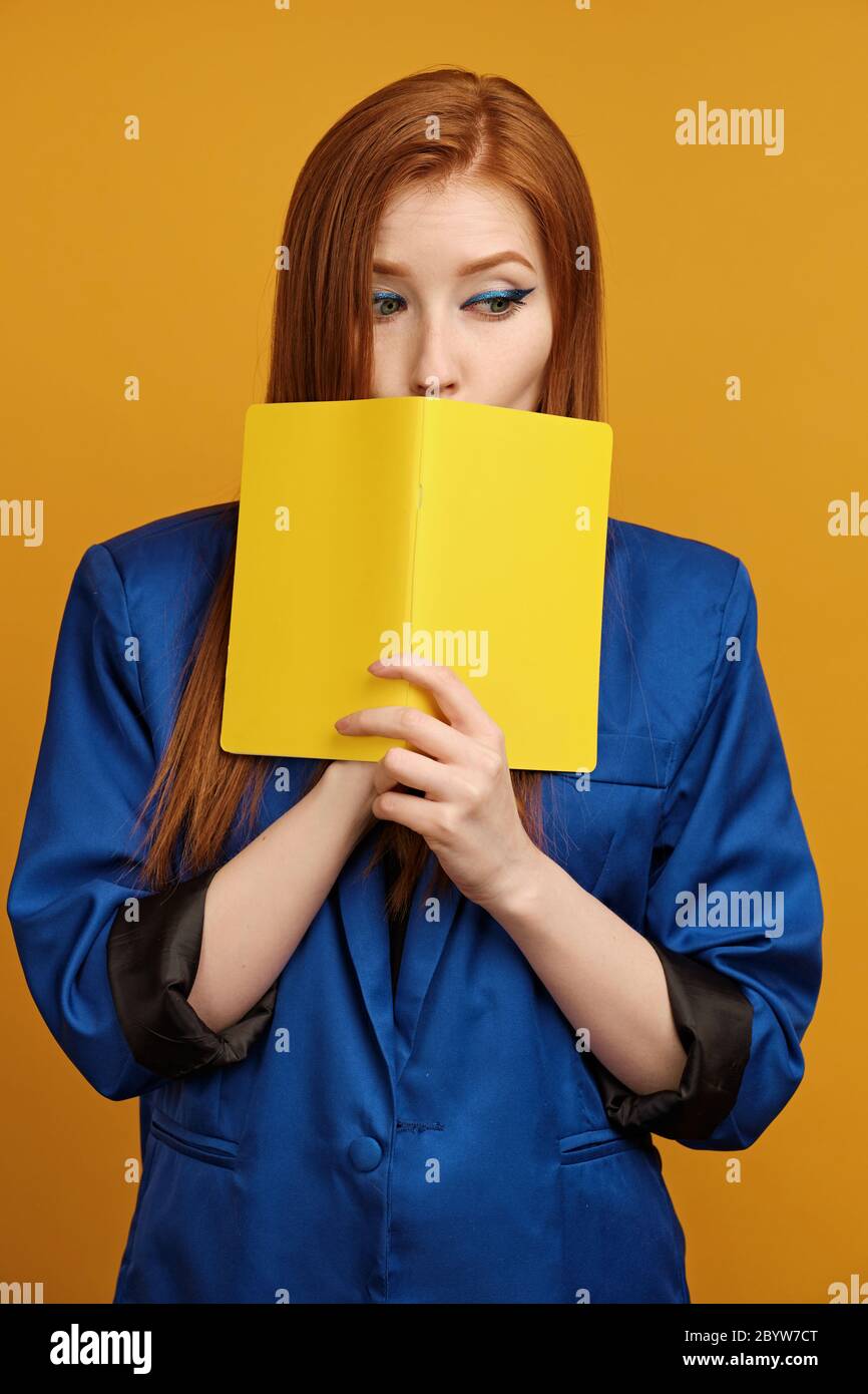 Redhead girl in a blue jacket stands on a yellow background, covering mouth with a yellow notebook and looks with eyes to the side Stock Photo