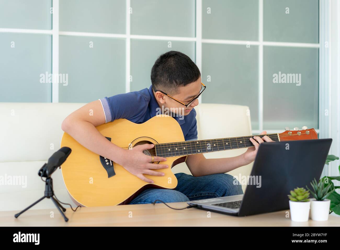 Asian boy playing acoustic guitar and watching online course on laptop while practicing at home. Asian boy virtual happy hour meeting for play music o Stock Photo
