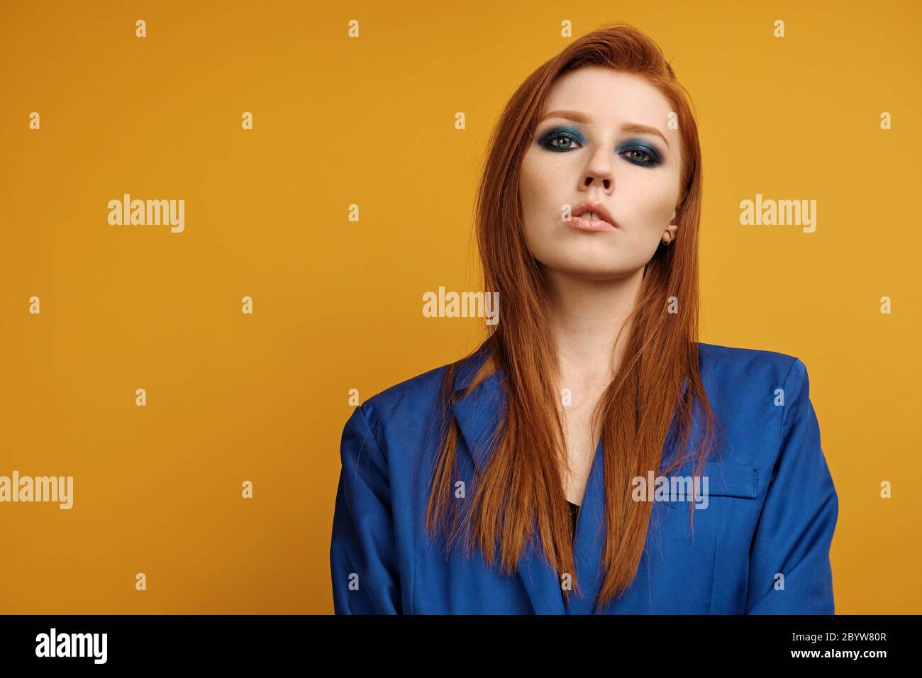 Headshot. Red-haired girl in a blue jacket and with blue eye make-up stands on a yellow background and looks in the frame Stock Photo