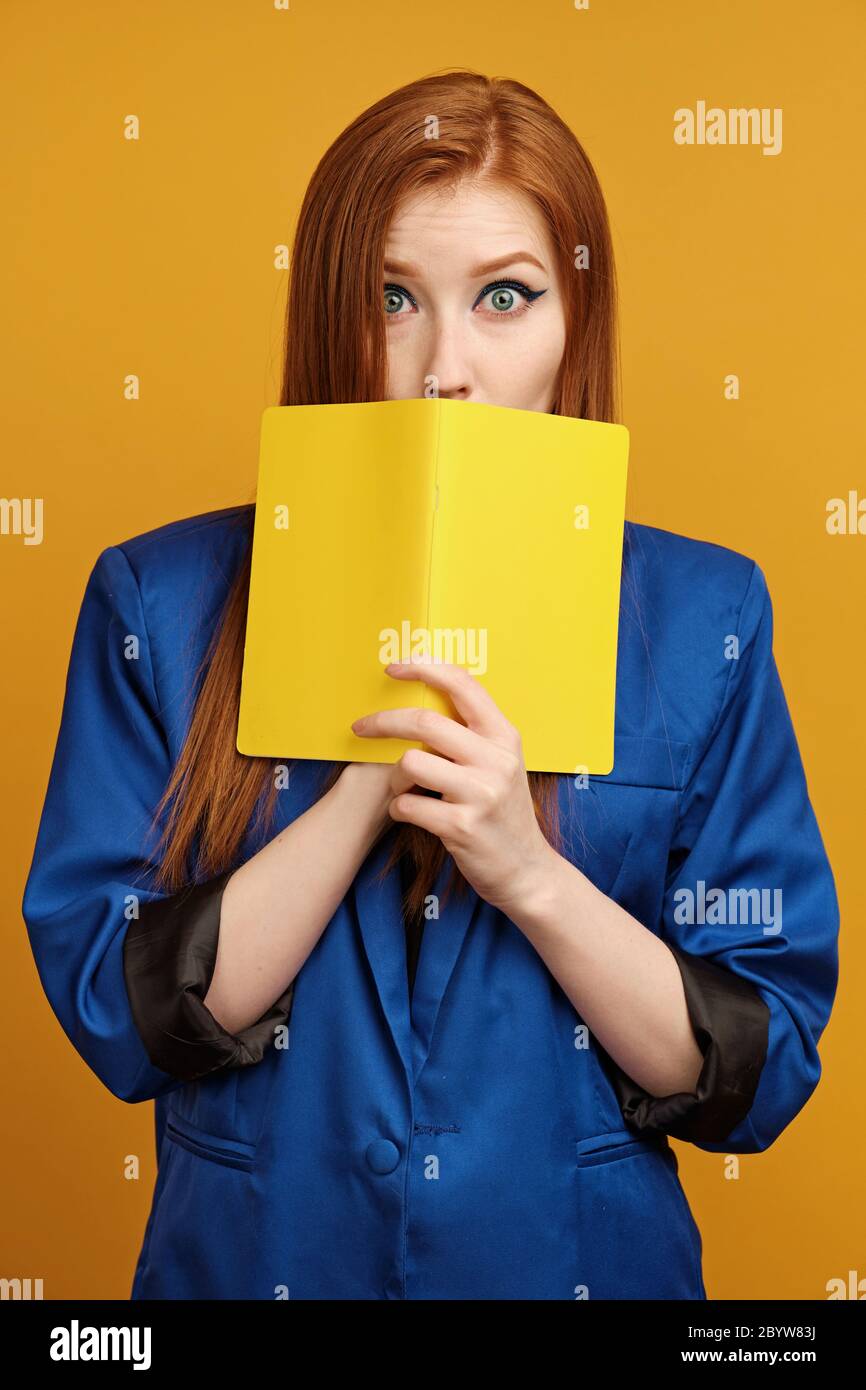 Red-haired girl in a blue jacket stands on a yellow background with his eyes wide open and covering mouth with a yellow notebook Stock Photo