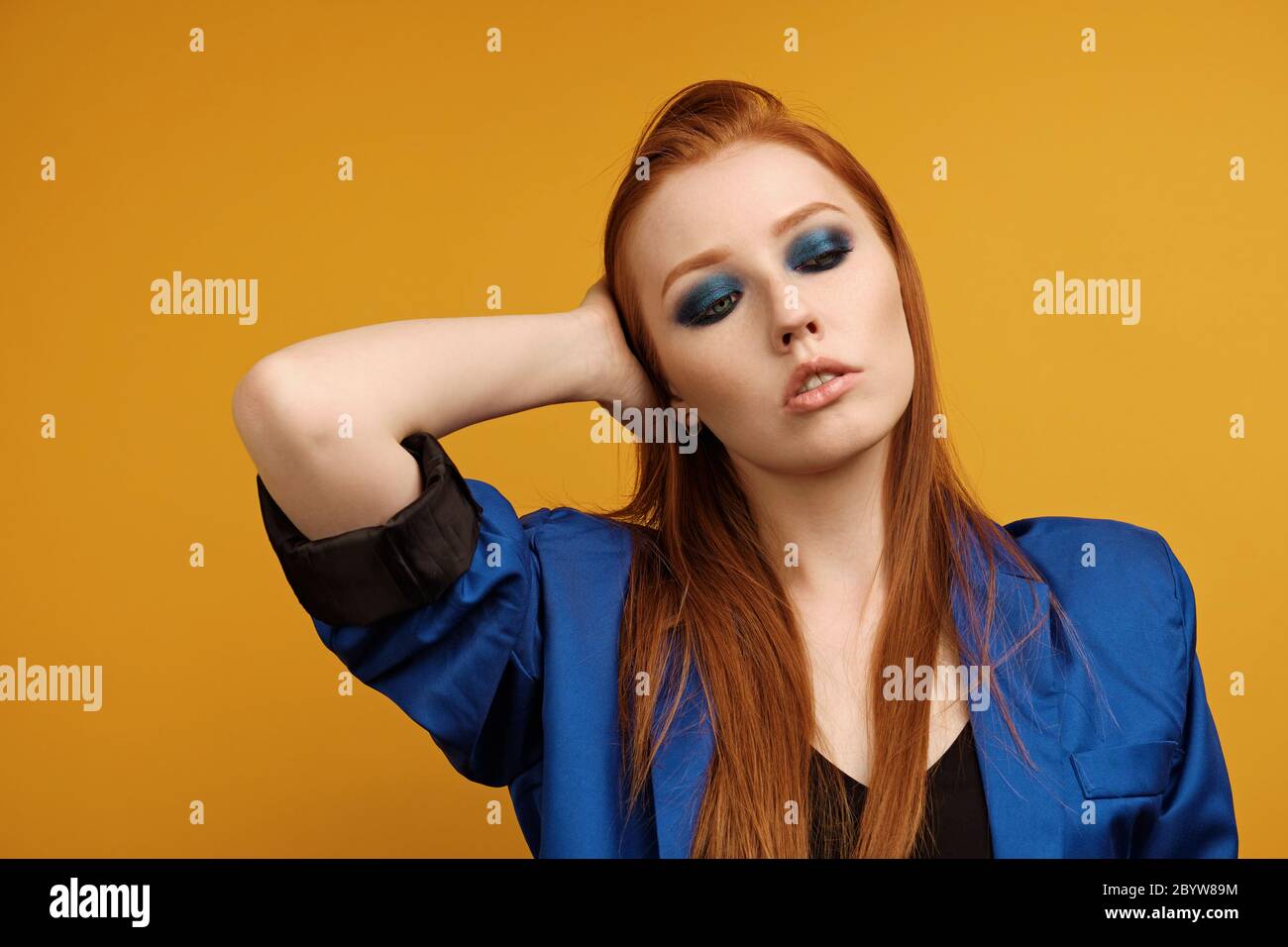 Redhead girl in a jacket and with blue eye makeup is standing on a yellow background, running hand through hair and looking down Stock Photo