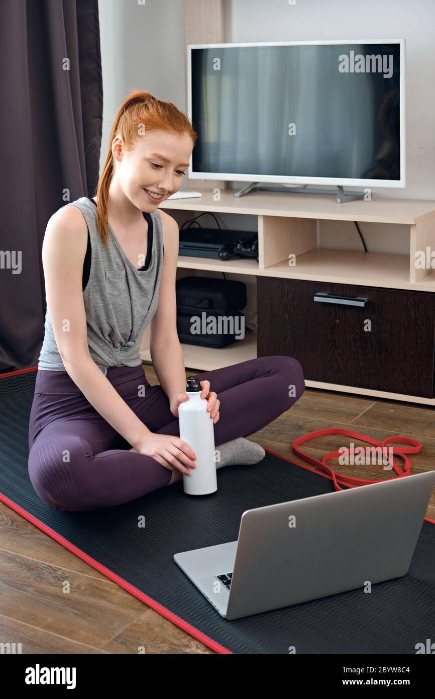 Redhead girl in a sports uniform sits on a mat with a bottle of water on the floor at home and looks at the laptop with a smile Stock Photo