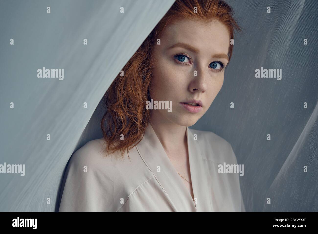 A red-haired girl with blue eyes stands against a white translucent curtain and looks into the frame. Stock Photo
