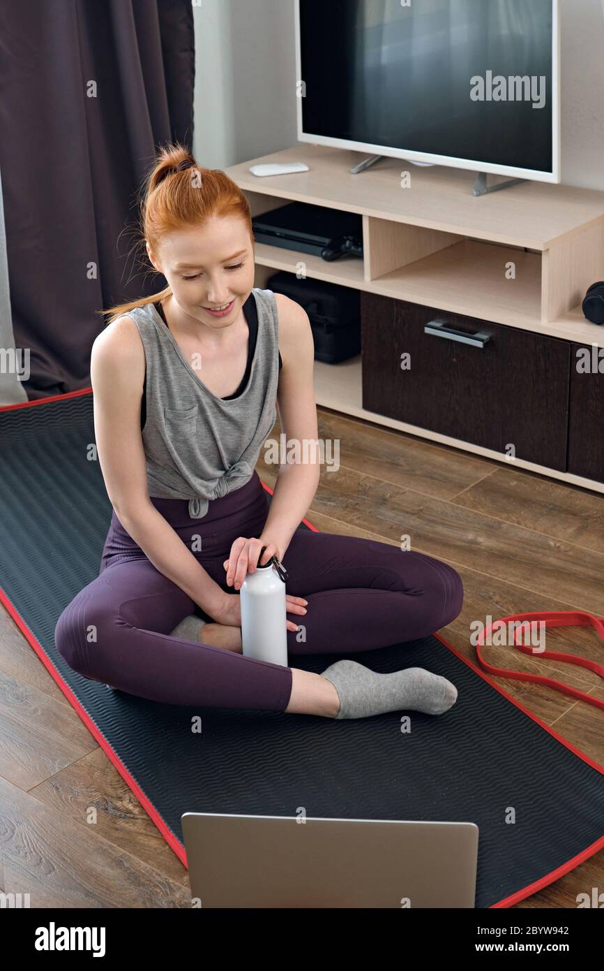 Red-haired girl in a sports uniform sits on a mat on the floor at home and looks into a laptop with a smile, frame from above Stock Photo