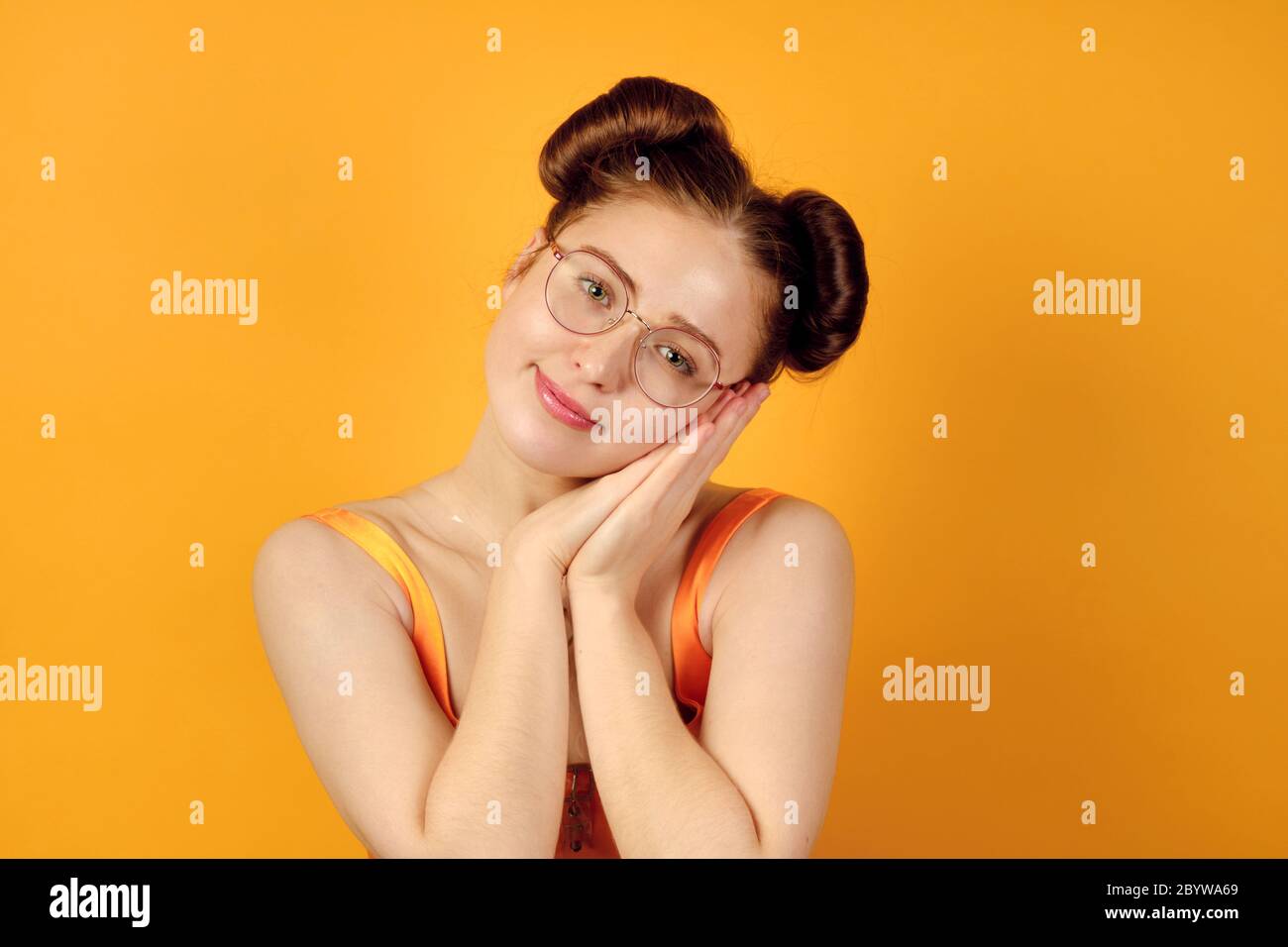 Red-haired girl in round glasses and an orange top stands on a yellow background, resting her head on folded palms. Stock Photo