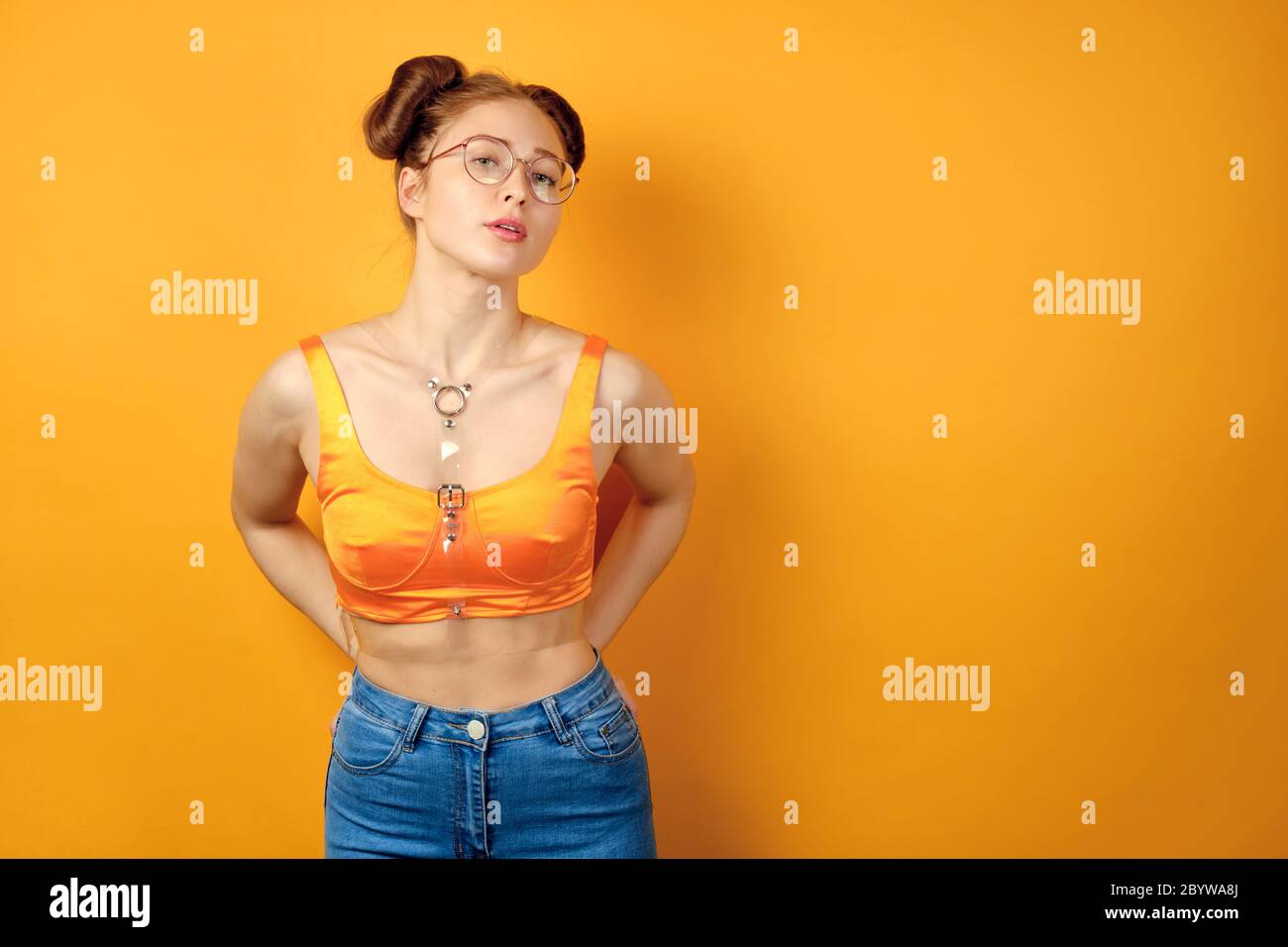 Redhead girl in round glasses and an orange top stands on a yellow background, holding her hands in the back pockets of jeans Stock Photo