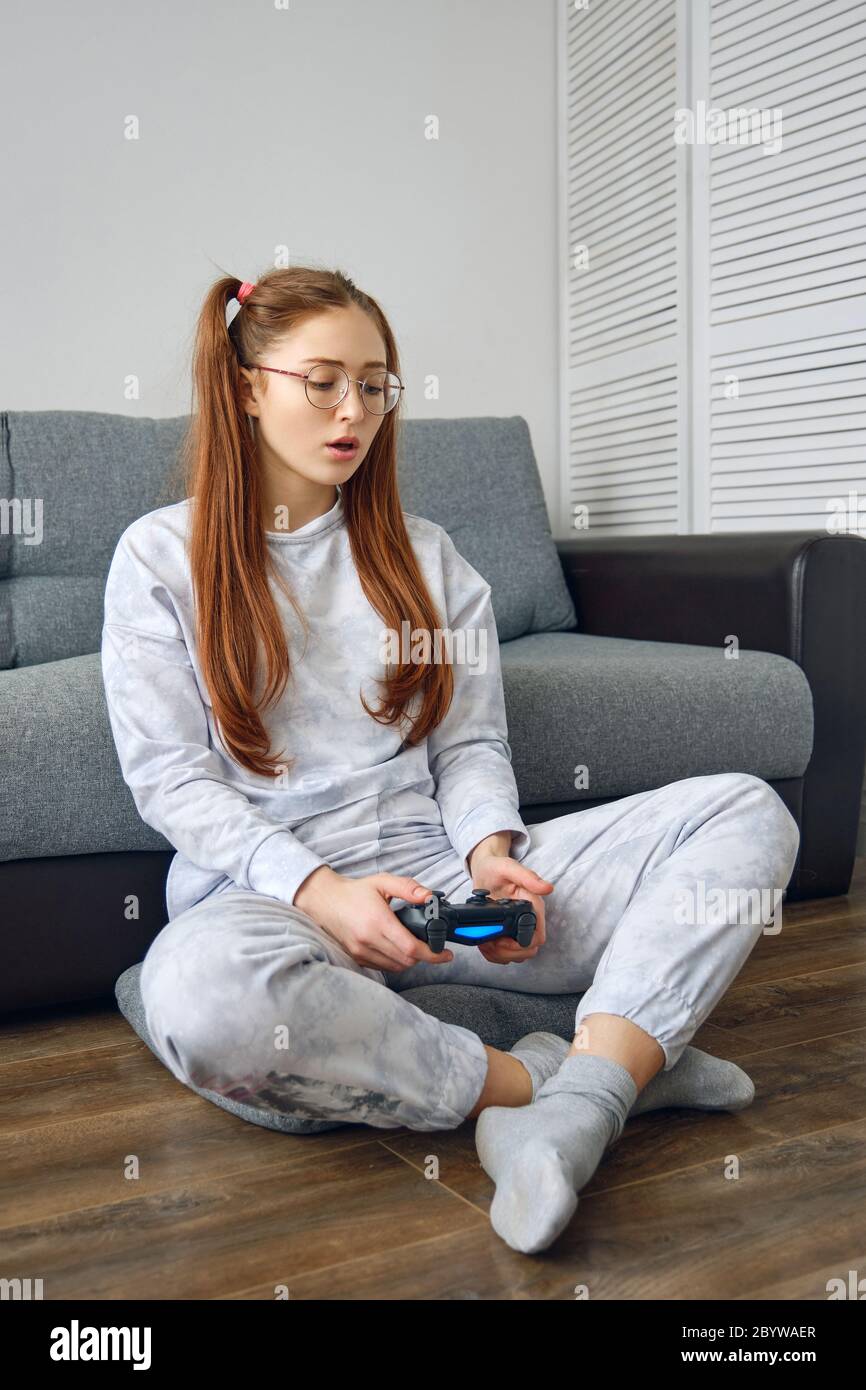 Redhead girl in pajamas and round glasses sits on the floor in front of the sofa with a game joystick in her hands and looks down. Stock Photo