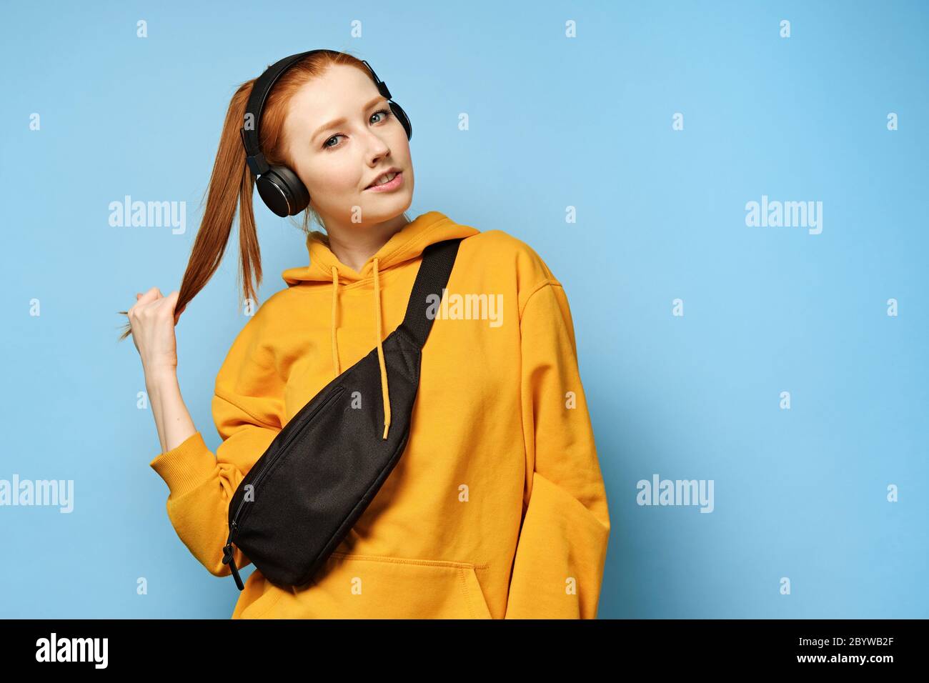 Red-haired girl in a yellow sweatshirt and with a black bag stands on a blue background, holding her tail with her hand Stock Photo