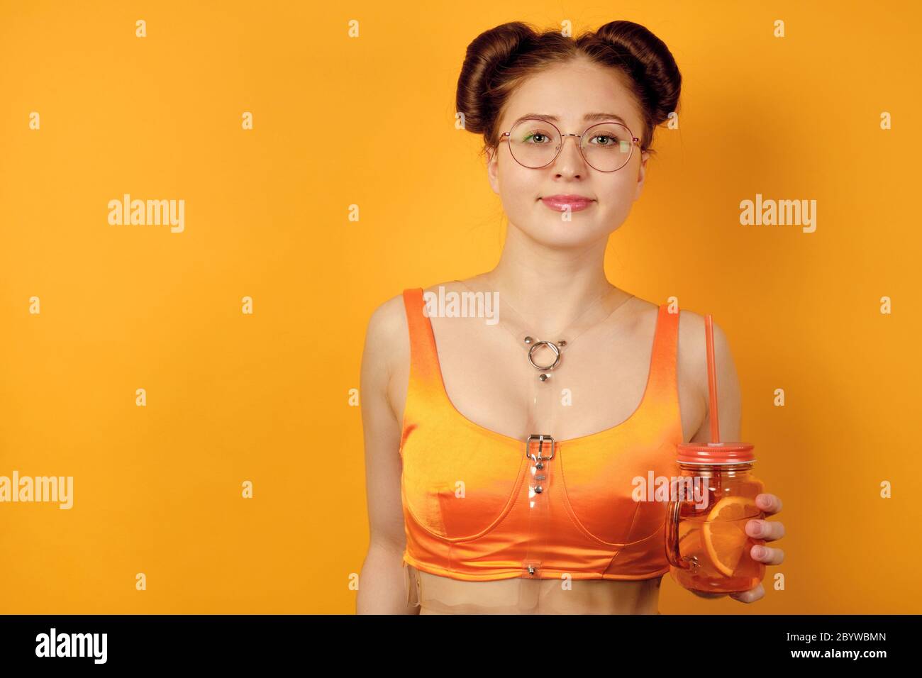 Redhead girl in round glasses and an orange top is standing on a yellow background with a glass of lemonade with a straw in hands Stock Photo