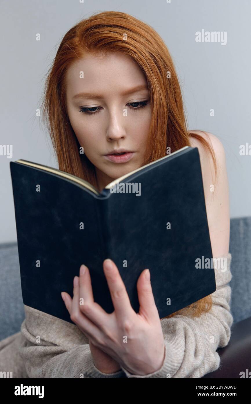 Red-haired girl sitting on the couch and thoughtfully reading a black book in her hands. Stock Photo