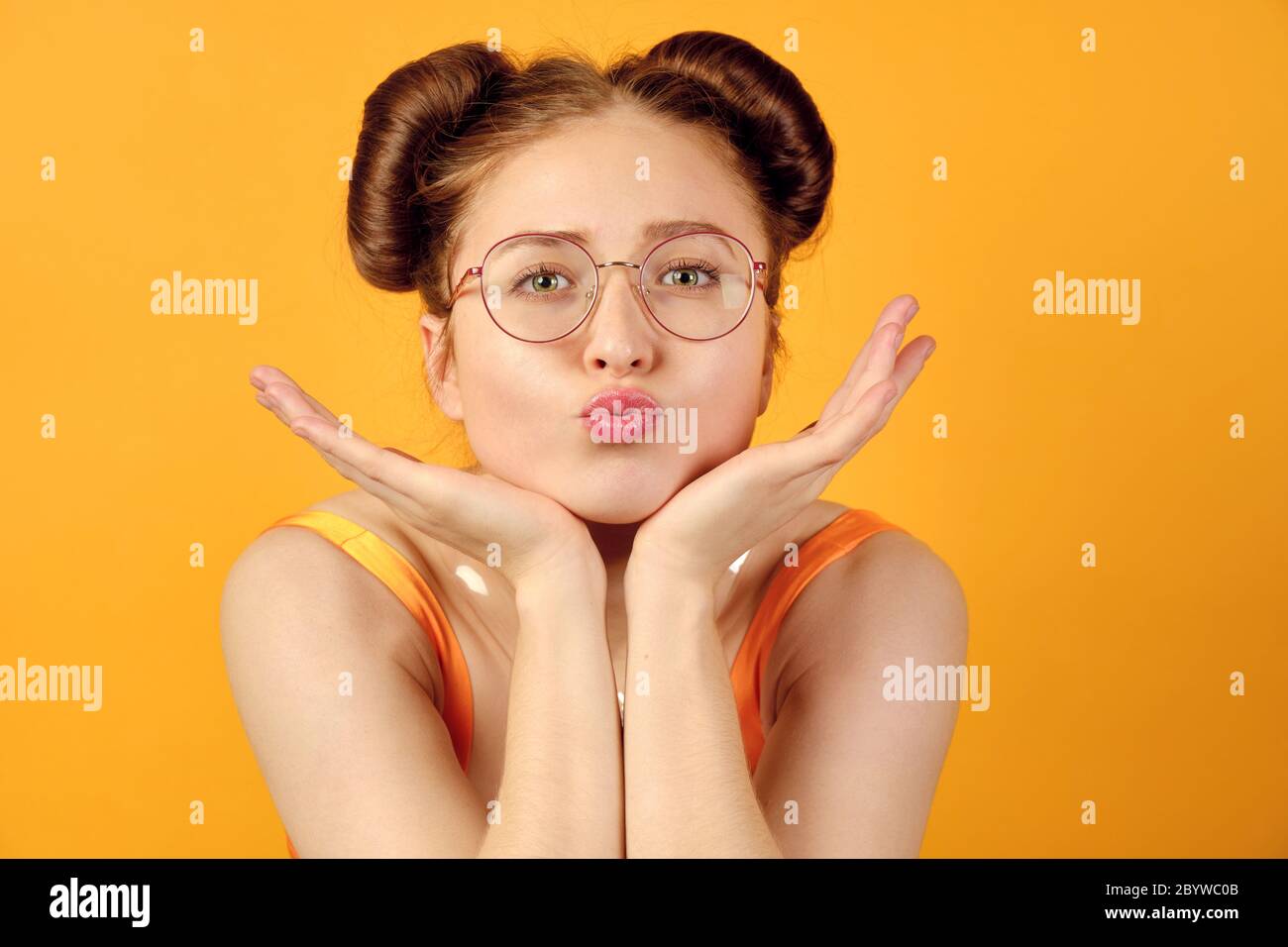 Red-haired girl in round glasses and an orange top is standing on a yellow background, chin resting on palm, lips extended forward Stock Photo
