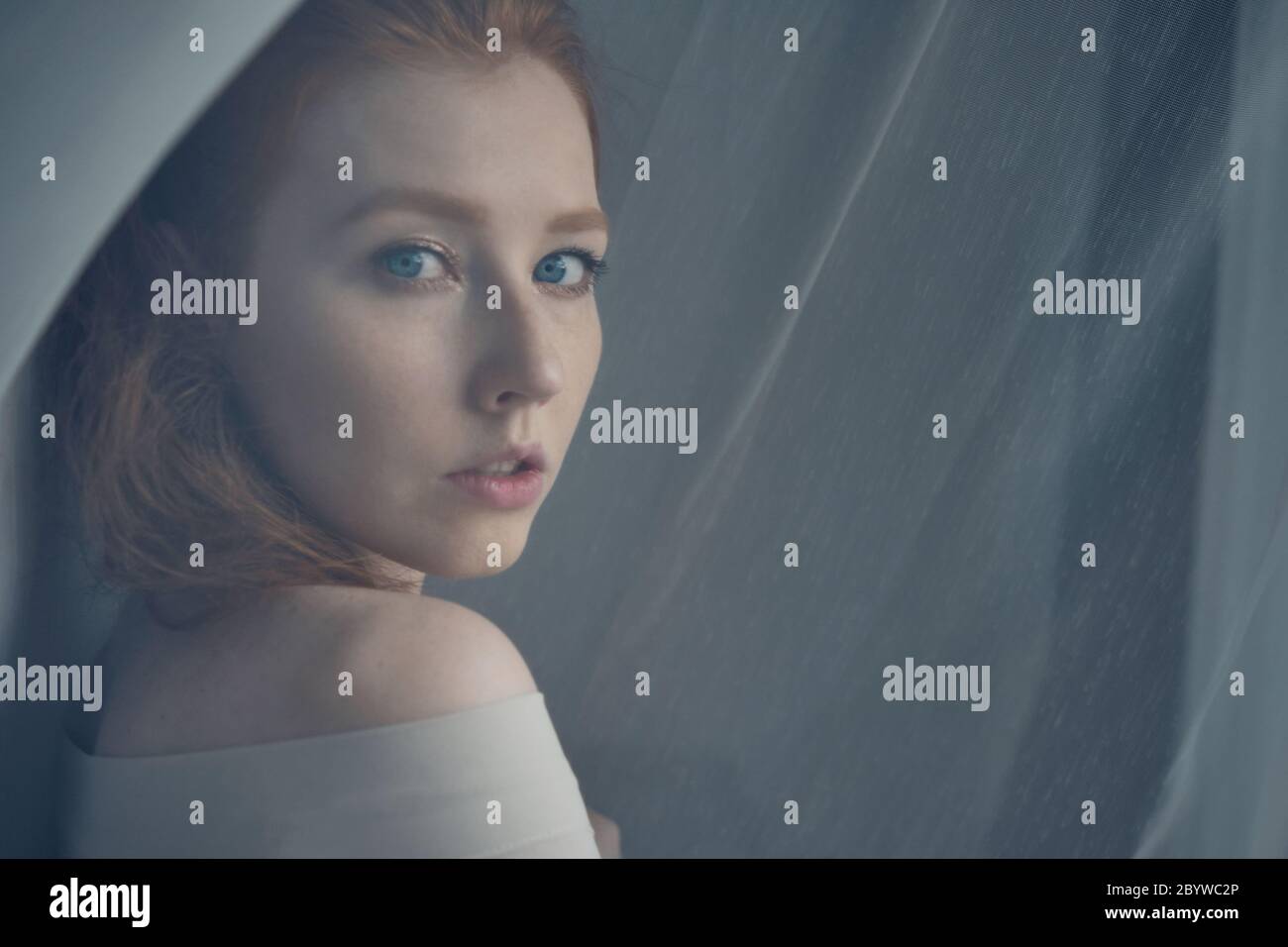 A red-haired girl with blue eyes stands in the midst of a translucent curtain and looks over shoulder, a view through the curtain. Stock Photo
