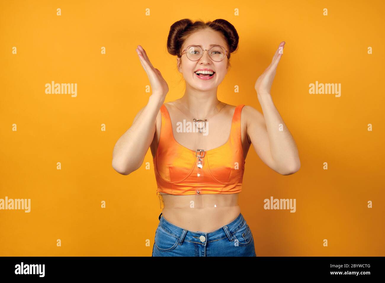 Redhead girl in round glasses and an orange top stands on a yellow background, laughs, raising palms up Stock Photo