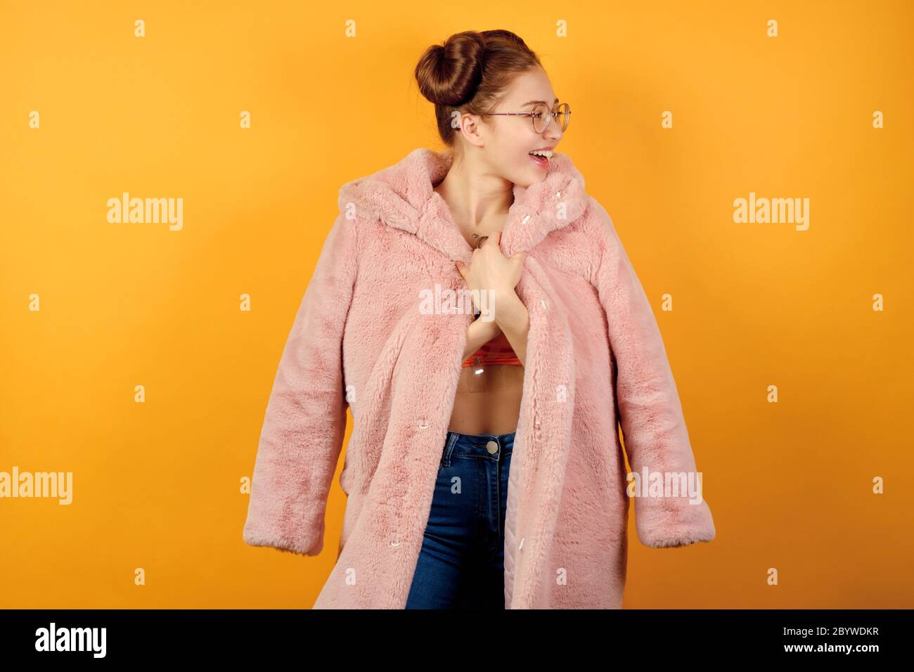 Redhead girl in glasses stands on a yellow background, looks joyfully to the side, adorning the edges of a draped pink fur coat Stock Photo