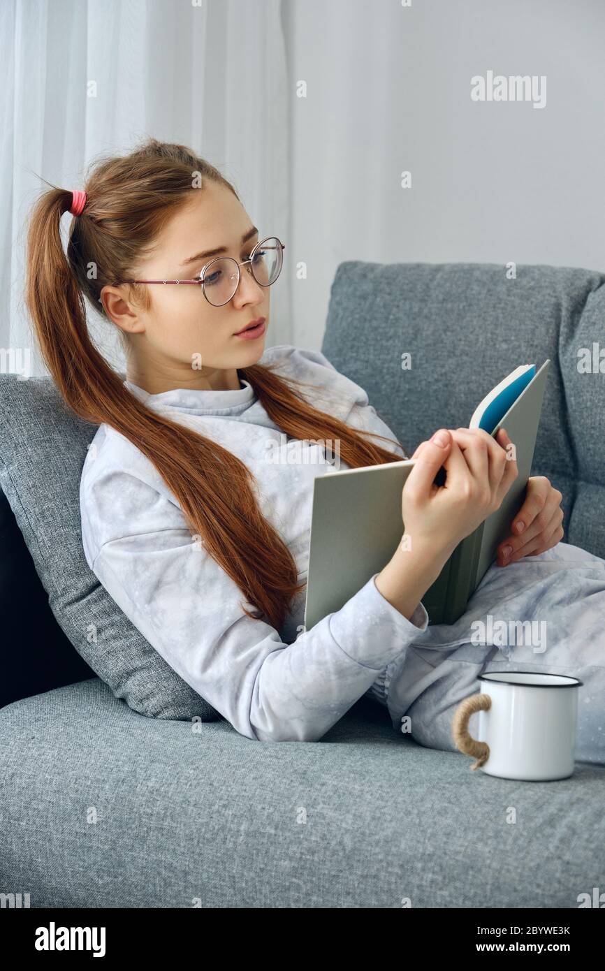Redhead girl in pajamas and round glasses lies on the sofa and reads a book. Stock Photo