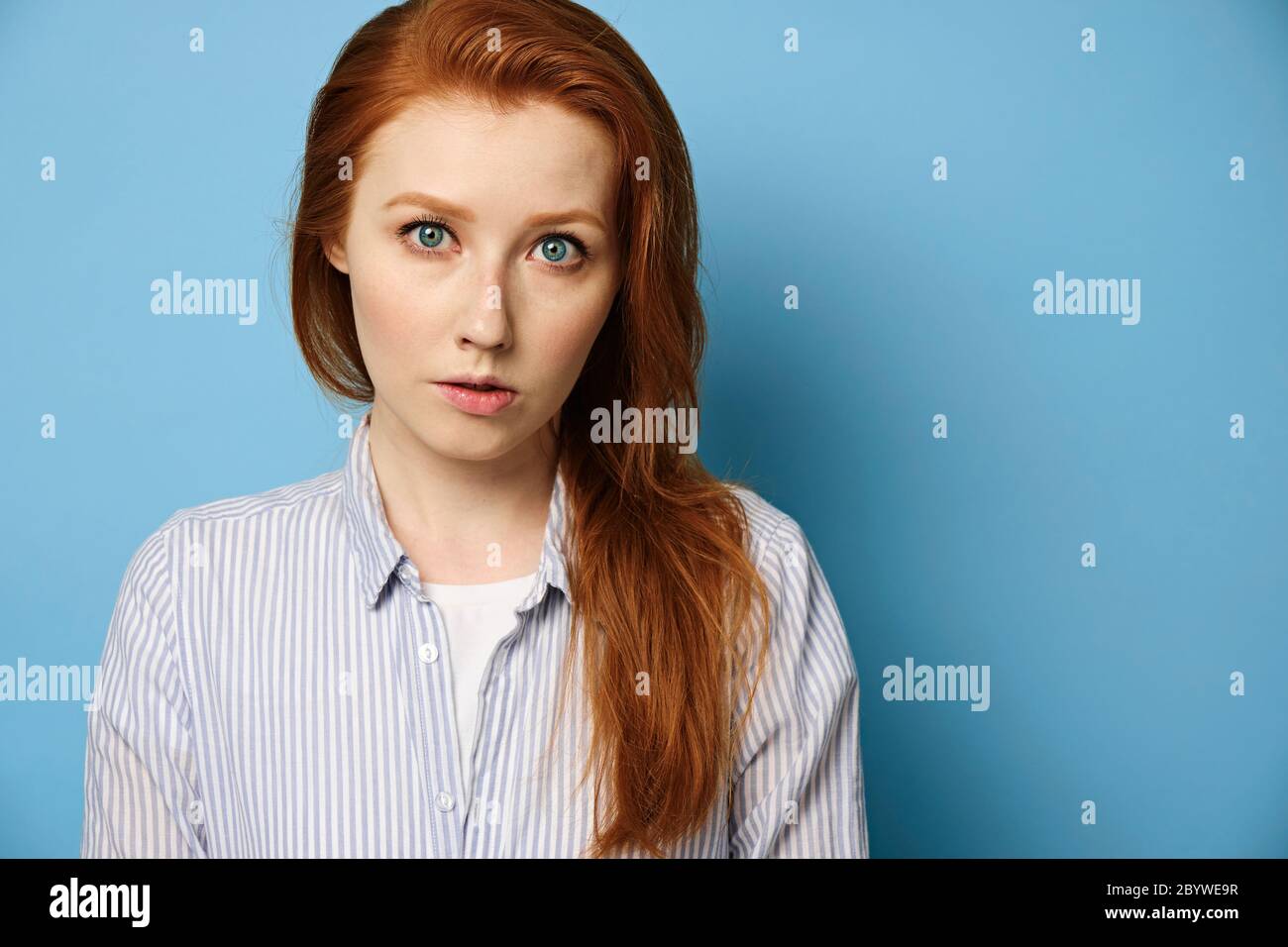 Red-haired girl with blue eyes in a striped shirt stands on a blue background and looks in the frame. Stock Photo