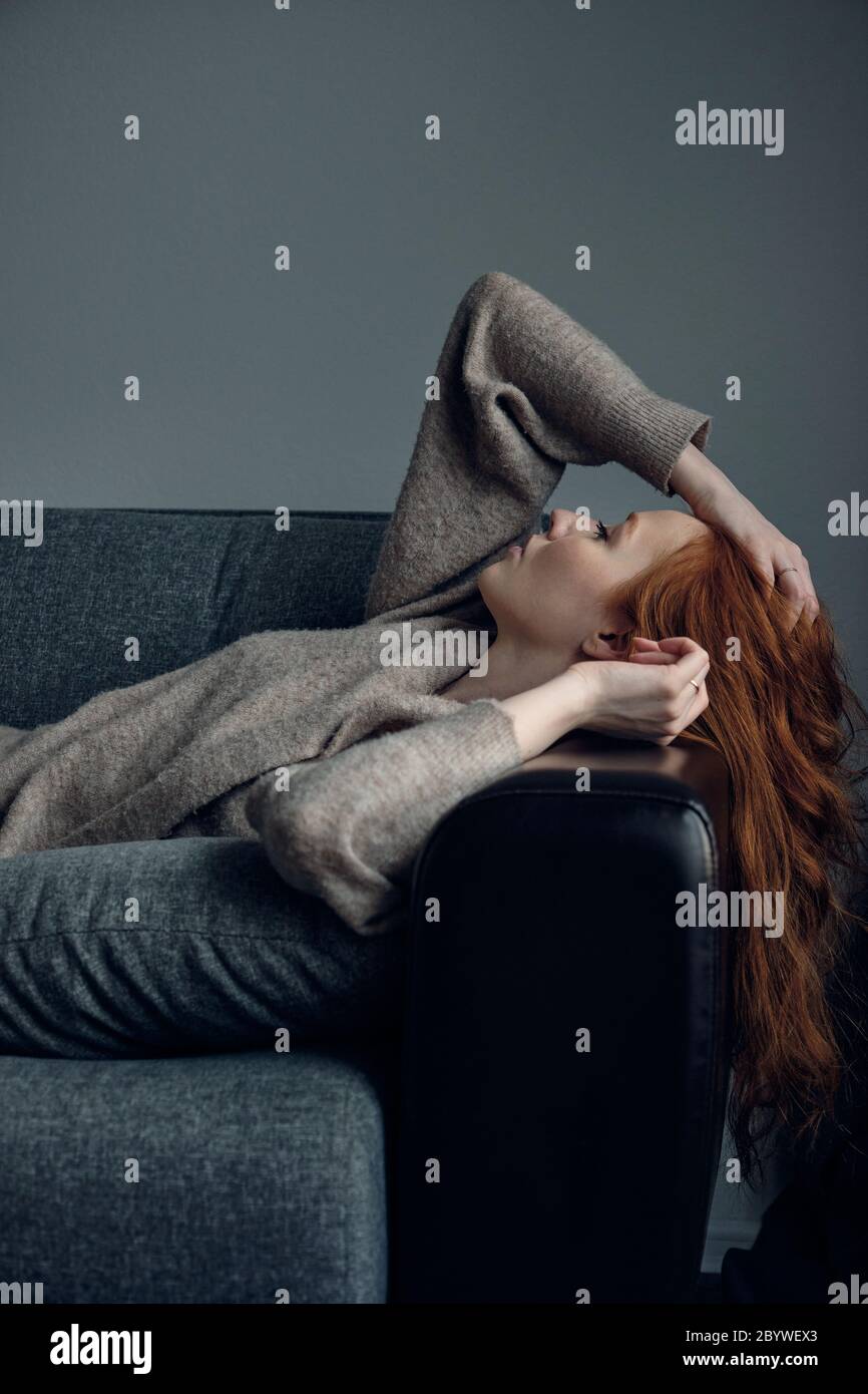 A red-haired girl in a beige cardigan lies on the sofa, leaning her head back on the armrest, running her hand through her hair. Stock Photo
